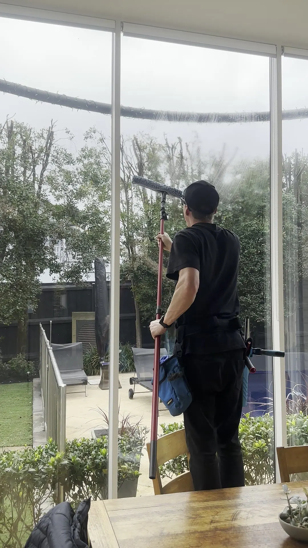 Cleaner squeegeeing the top of a floor-to-ceiling glass wall from inside using a pole, garden visible through the glass