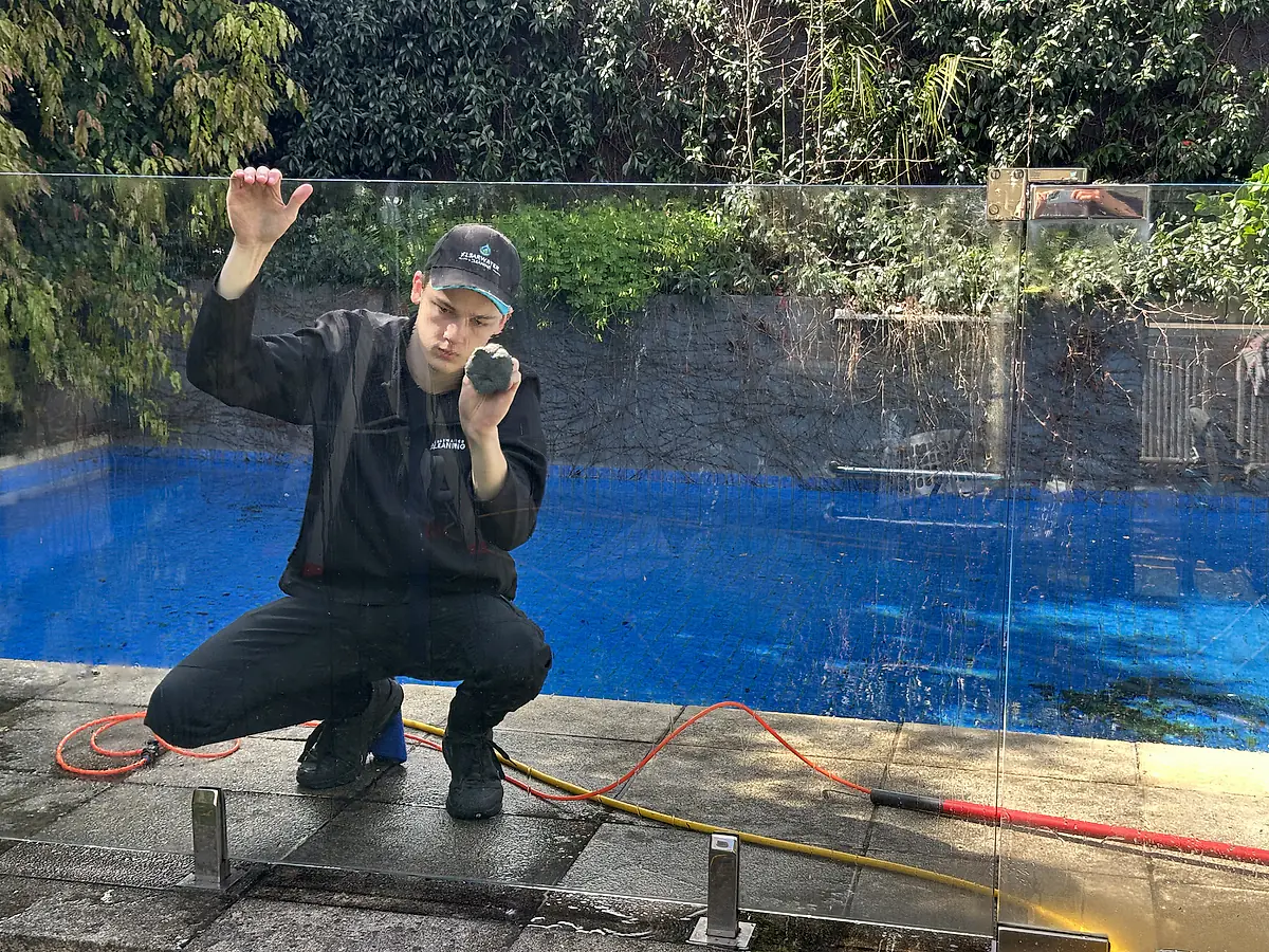 View through a cleaned glass pool fence panel to a Clearwater cleaner working on the far side, pool water visible through the glass